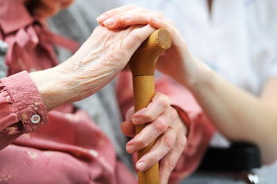Die Hand einer Pflegerin liegt auf der Hand einer älteren Frau, die wiederum einen Stock umfasst